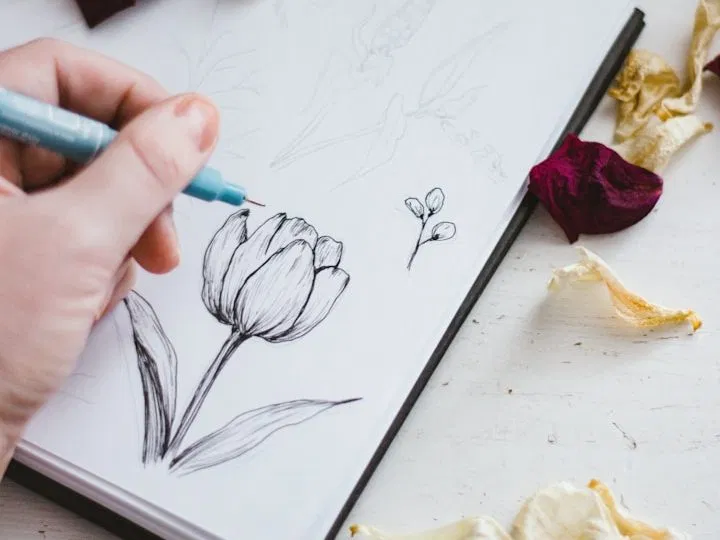 A hand sketching a flower on a desk, representing the creative process as a spiritual practice.